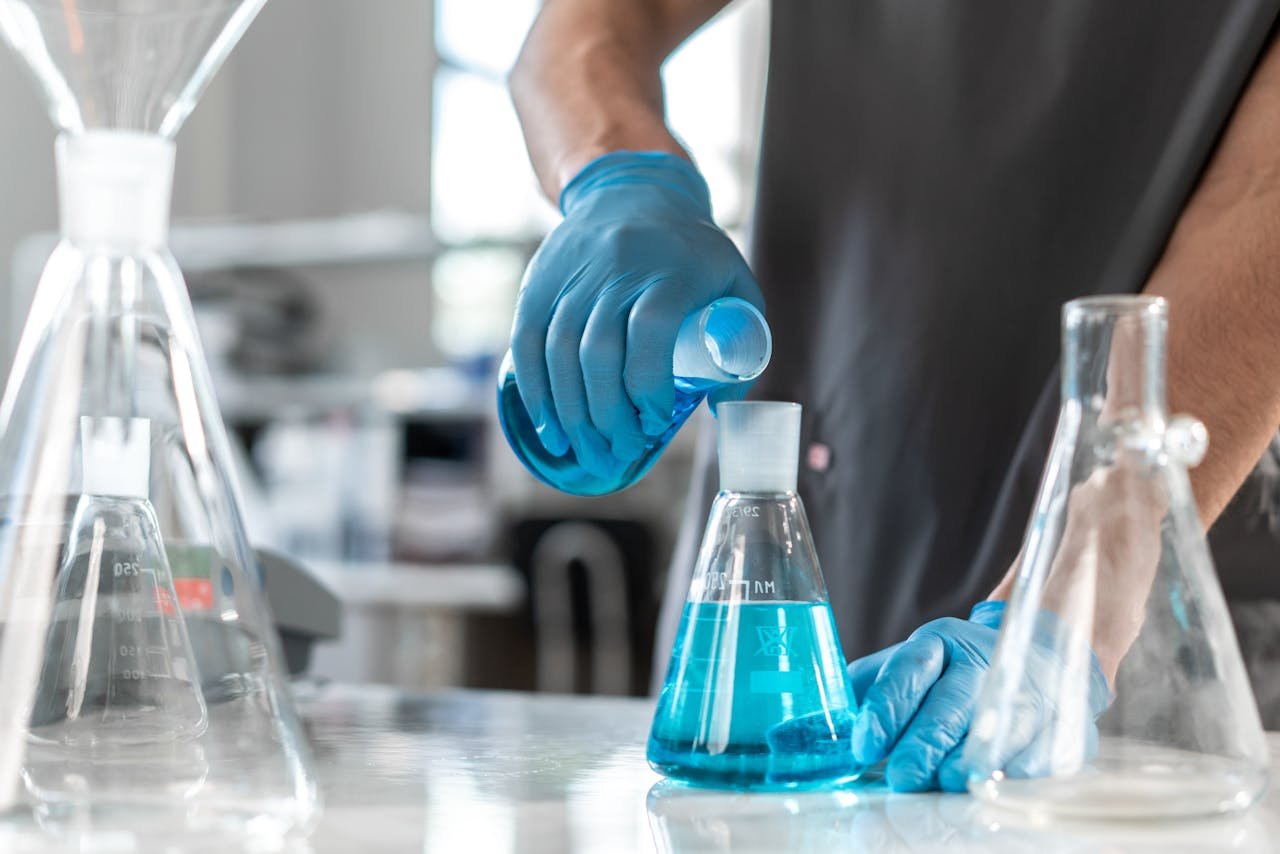 Scientist wearing gloves pours blue liquid into an Erlenmeyer flask in a laboratory setting.